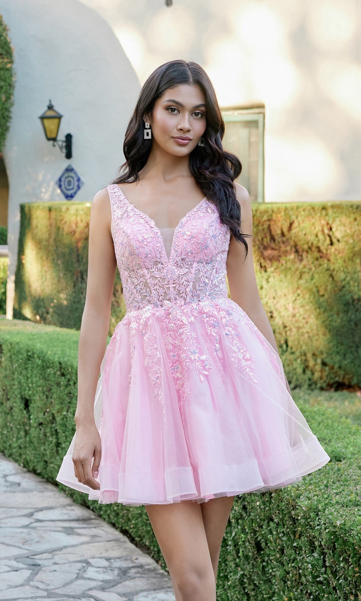 A woman stands outdoors surrounded by hedges and a sunlit garden path, wearing the Juliet JT987N Short Homecoming Dress in blush—a sleeveless design with a lace-embroidered bodice and tulle skirt.